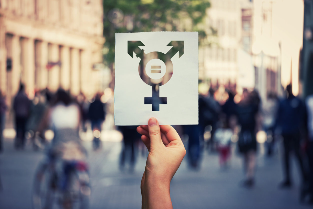 Une main tient un carton sur lequel apparaît un symbole transgenre. les symboles du féminin et du masculin sont liés dans un même dessin