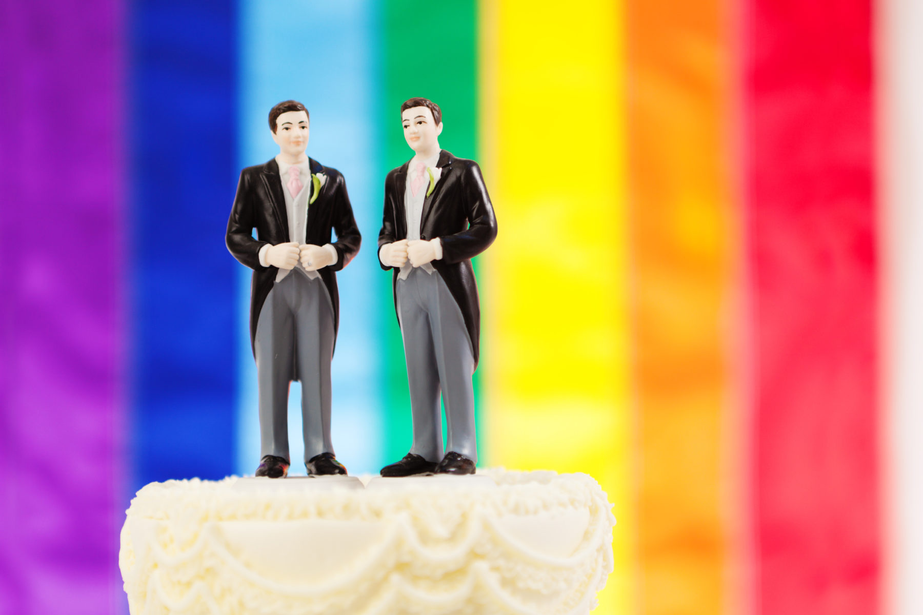 Deux petites figurines d'hommes en costumes au sommet d'un gâteau de mariage. En arrière-plan, les couleurs du drapeau LGBT, arc-en-ciel