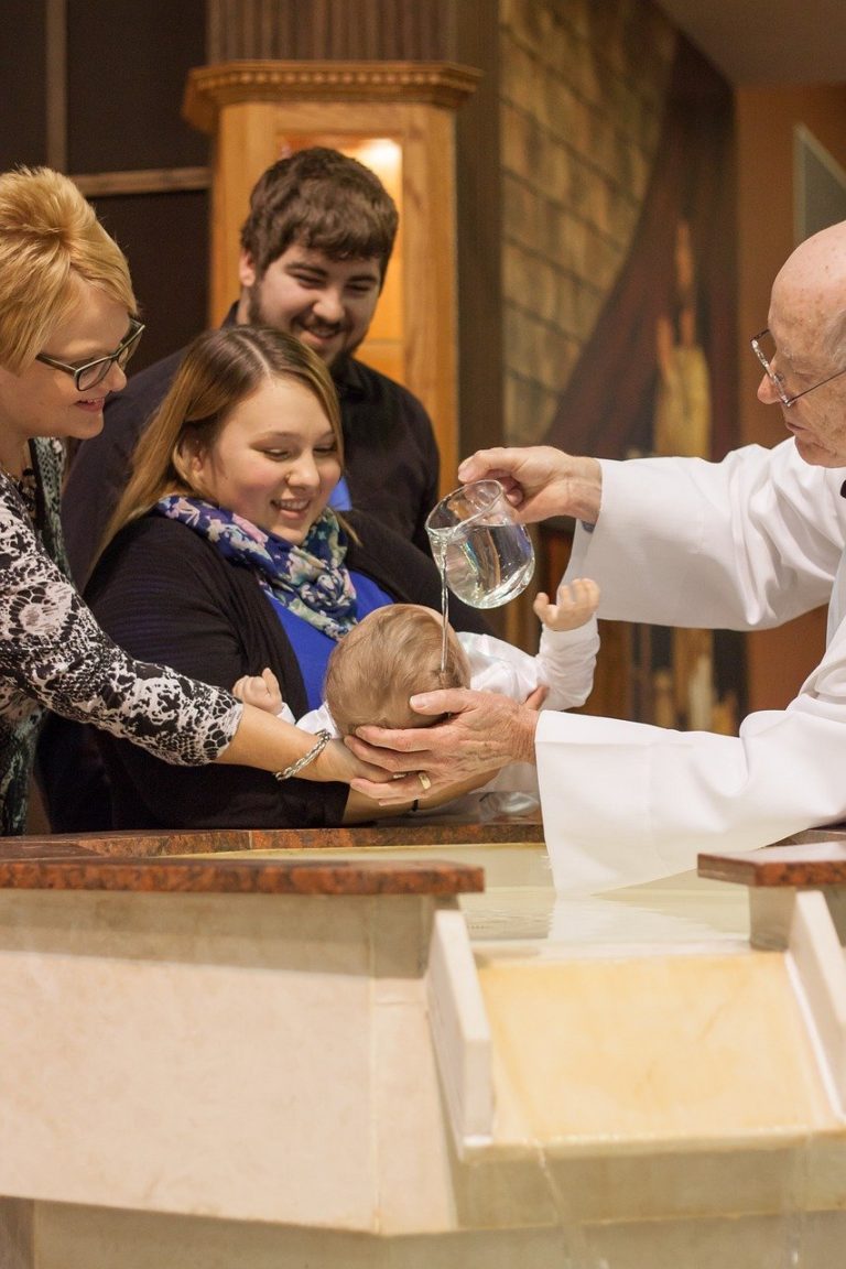 Cérémonie d'un baptême catholique: un prêtre verse de l'eau sur la tête d'un nourisson, en présence de trois membres de la familles, souriants