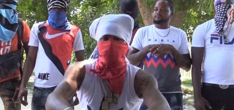 Un membre du gang haïtien "400 mawozo" se tient accroupi au premier plan, la tête et le bas du visage cachés par des foulards. Derrière lui, d'autres membres du gang, debout