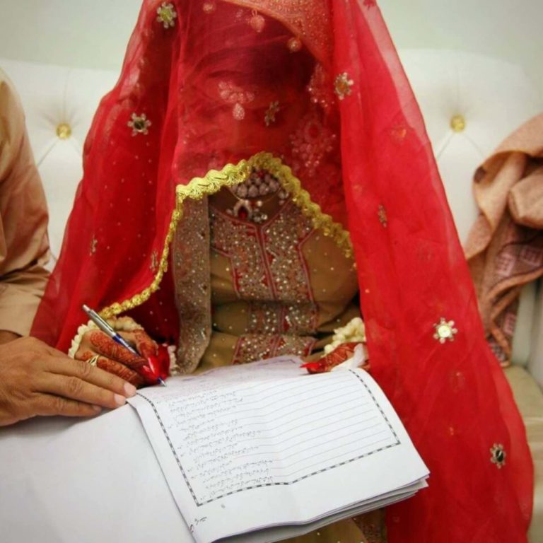 Une jeune mariée pakistanaise signe des documents de mariage. Elle est penchée en avant et couverte d'un voile rouge, on ne voit pas son visage