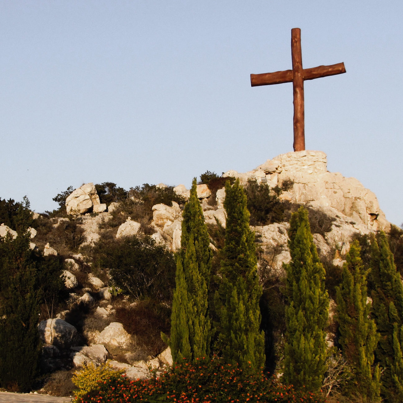 Une croix en bois surmonte une colline rocheuse, sur fond de ciel bleu. La colline est parsemée d'arbres.