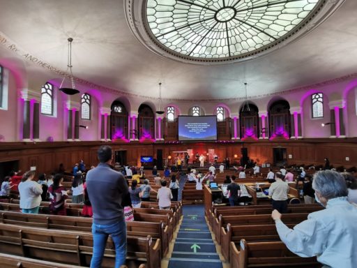 Panorama de l'intérieur d'une vaste église avec des bancs en bois. De dos à l'objectif, les chrétiens regardent vers un grand écran qui affiche un chant.