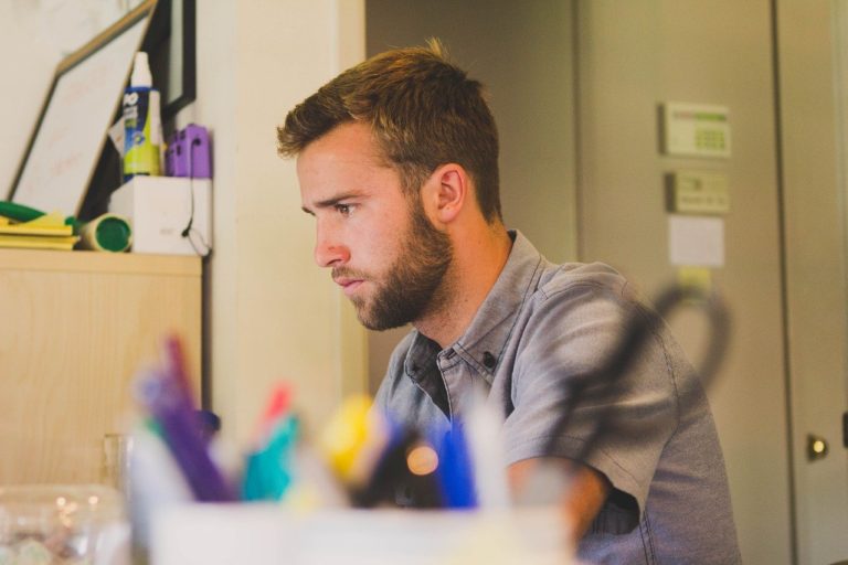 On voit la tête et les épaules d'un homme brun de profil, concentré sur son ordinateur. Au premier plan, un pot rempli de crayon, flou