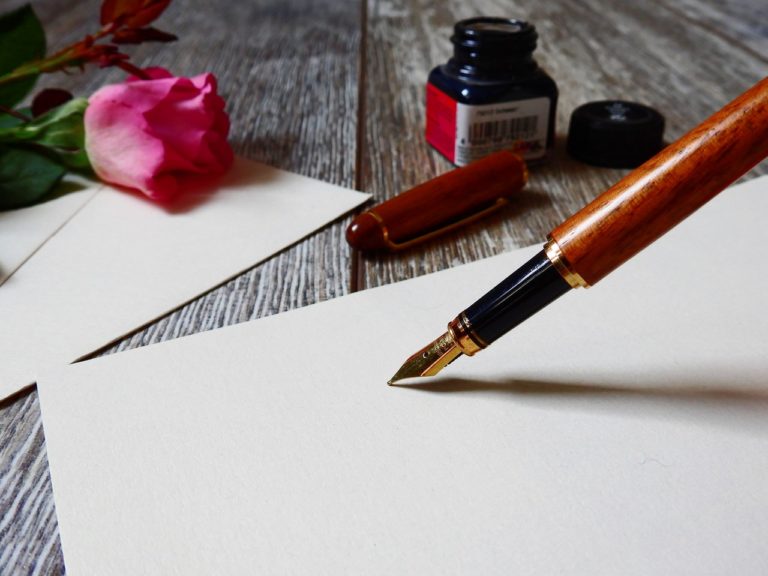 La pointe d'un stylo plume est posée, prête à écrire, sur une feuille blanche. A côté se trouvent un encrier et une rose rose