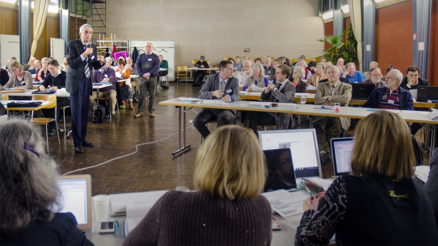 Synode de l'EERV en 2019. Un homme parle dans un micro parmi des dizaines de participants, assis à des pupitres. Ils écoutent, écrivent ou parlent entre eux