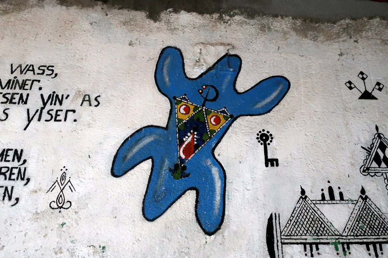 Sur un grand mur blanc, des graffitis écrits et des symboles. Une grosse forme bleue taggée, animale ou humaine, comporte des symboles en son centre