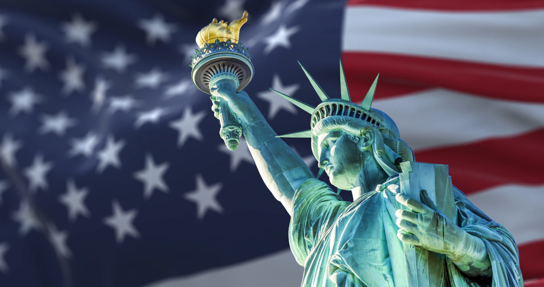 Vue en contre-plongée de la statue de la liberté devant le drapeau américain. Elle est bleue turquoise et le flambeau est doré
