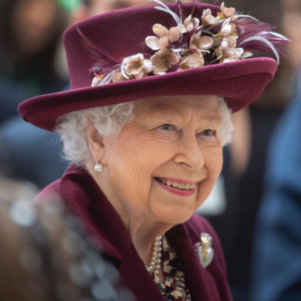 Photo portrait de la reine Elizabeth II, portant un chapeau rouge bordeaux fleuri. Elle sourit