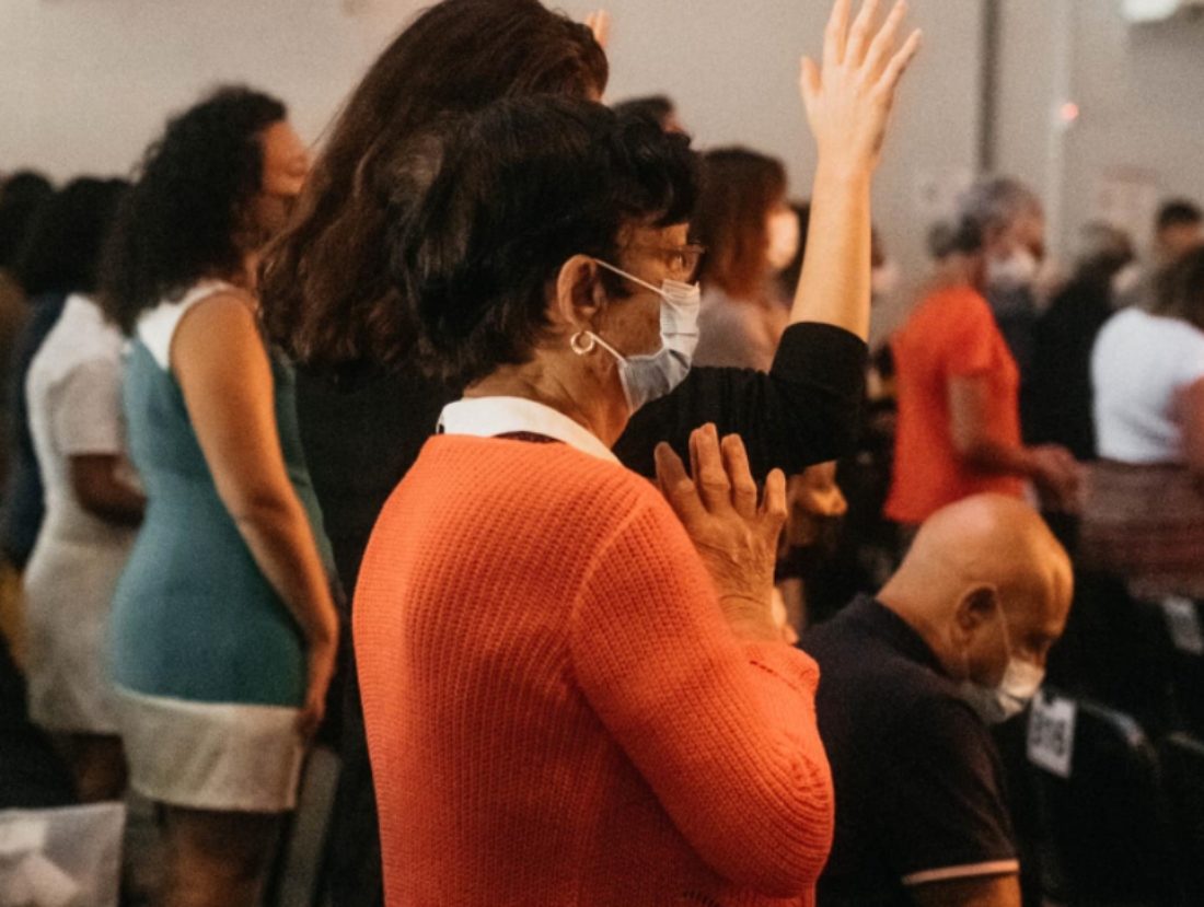 Des chrétiens debout dans une Eglise. Au premier plan, une dame habillée en orange, portant un masque anti-covid, a les mains jointes.