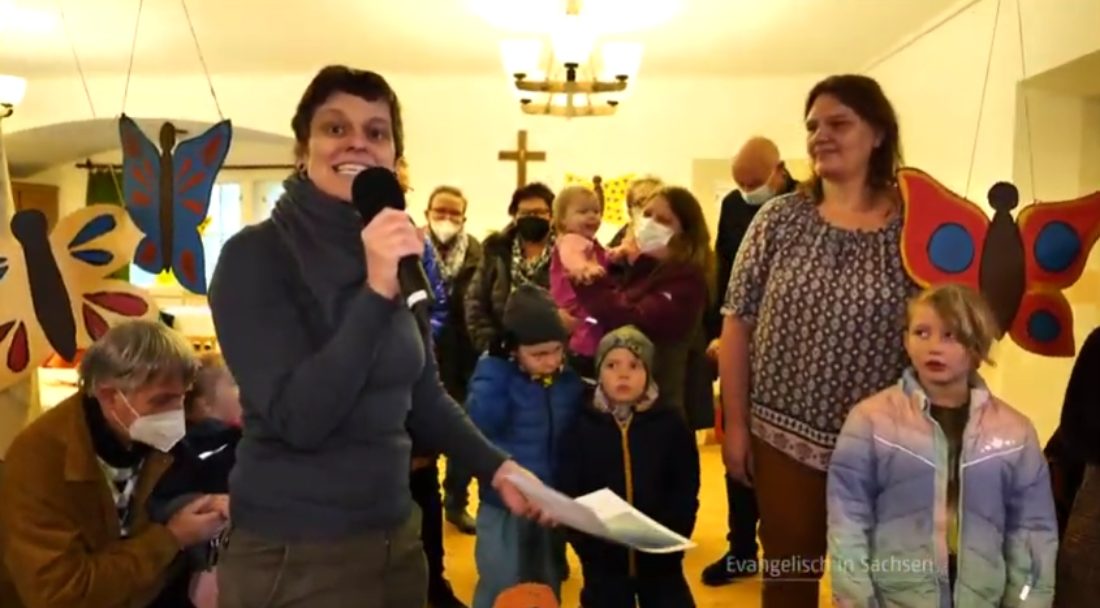 Une présentatrice au cheveux courts parle dans un micro, devant un groupe de personnes qui portent des masques anti-covid. Ils sont dans une petite salle éclairée à la lumière électrique.