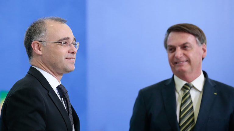 André Mendonça et Jair Bolsonaro, en costumes noirs, souriant. Derrière eux, un mur bleu uni.