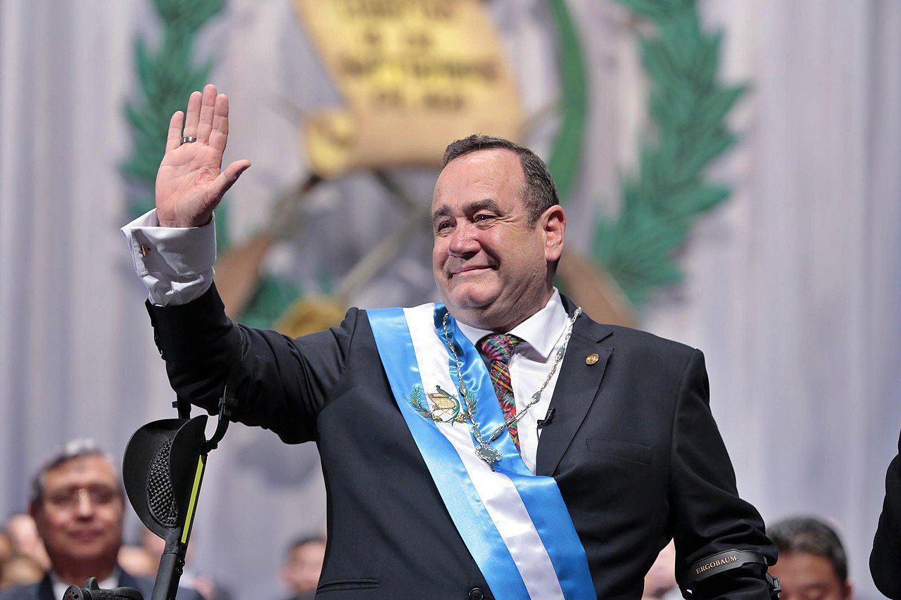 Alejandro Giammattei, environ 65 ans, lors d'un discours. Il sourit et lève le bras droit en signe de salutation. Il porte sur son buste l'écharpe présidentielle du Guatemala, rayée de bleu et blanc