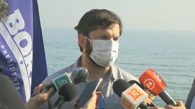 Gabriel boric, portant un masque anti-covid blanc, répond à la presse. Plusieurs micros sont tendus devant lui. En arrière plan, l'horizon de l'océan.