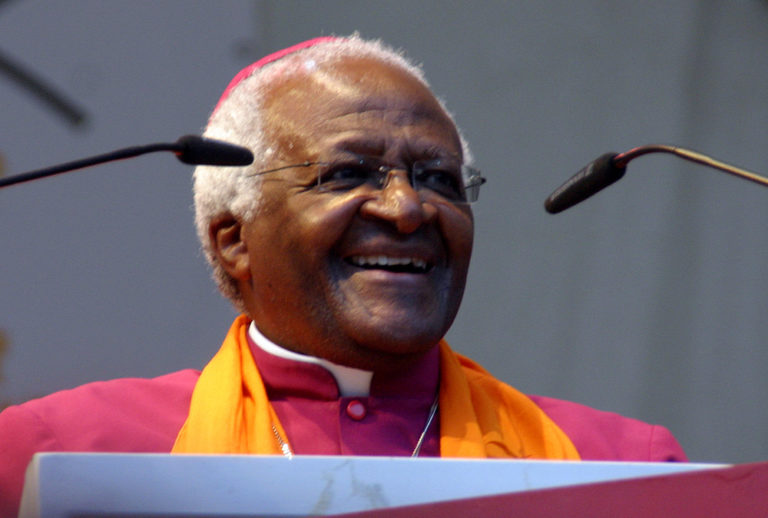 Desmond Tutu, homme âgé sud-africain, apparaît très souriant. Vêtu d'un habit d'évêque rose avec une écharpe orange, il parle derrières deux petits micros flexibles placés de part et d'autre de son visage.