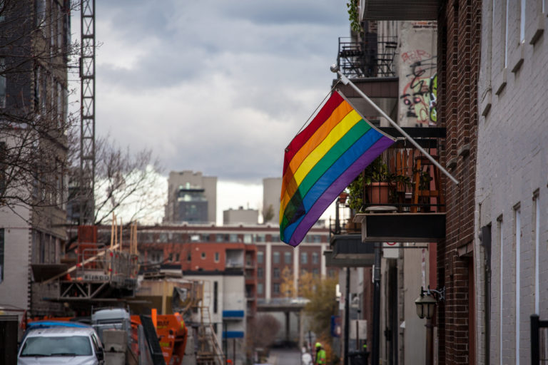 Dans une rue en pleine ville, un drapeau LGBT+, fixé sur un mur, vole au vent
