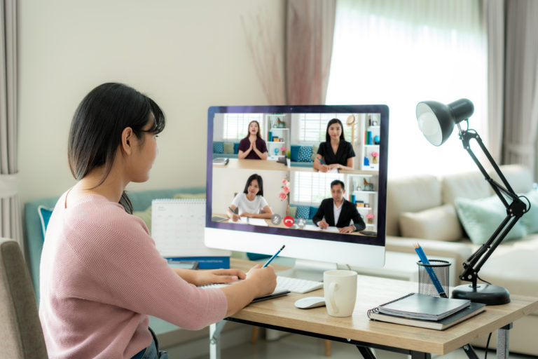 Une jeune femme asiatique, qui tourne presque le dos à l'objectif, est assise à son bureau; Elle est en visioconférence, et prend des notes.