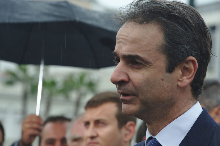 Kyriakos Mitsotakis, de profil, parle, entouré de personnes qui ne sont pas toutes dans le cadre. Près de lui, quelqu'un tient un parapluie. Il pleut.et l'atmosphère est grise.