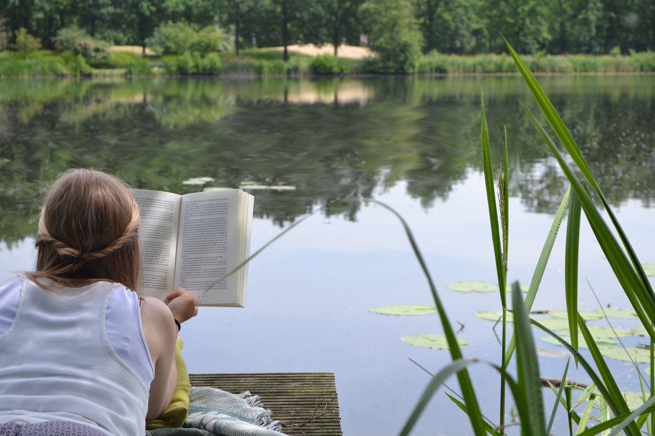 Une jeune femme, allongée sur un ponton donnant sur un étang, lit. On la voit de dos. Elle est sur le ventre et habillée en blanc.