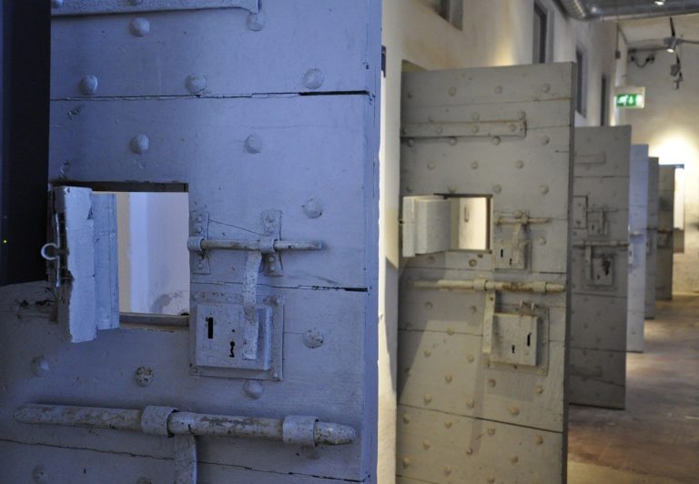 Plusieurs grosses portes blanches de prisons sont ouvertes, les unes à côté des autres. Vue de côté