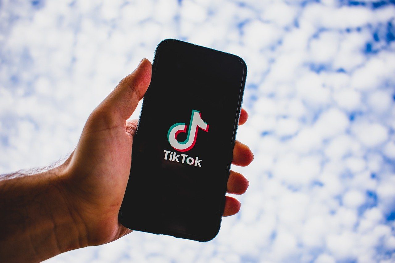 Une main tient un smartphone. Sur l'écran noir, le logo de Tik Tok apparaît en blanc. L'arrière plan est flou et blanchâtre.