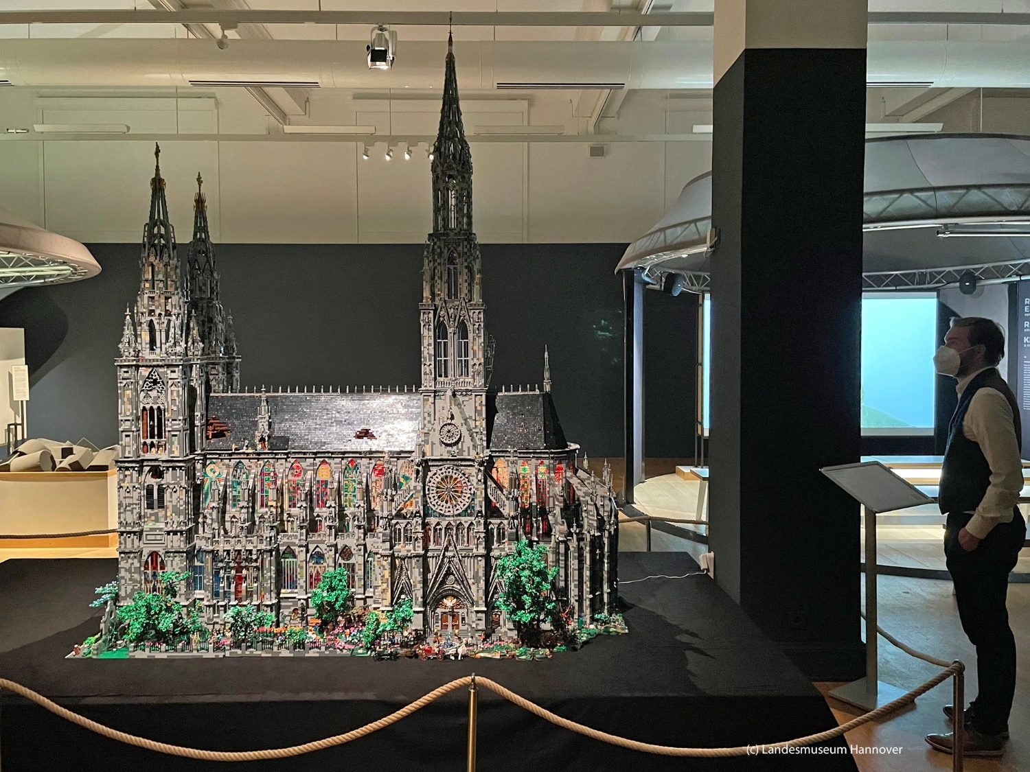 La "cathédrale Brickminster" dans une vaste salle du musée de Hanovre. La construction, haute de trois mètres, est posée sur un socle noir. On y perçoit une multitude de petit détails. Elle comporte trois clochers. A droite, un visiteur la contemple.