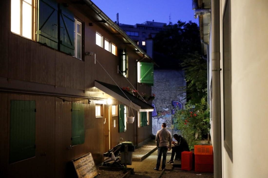 Vue du bâtiment de l'organisme le Passage, depuis la cour arrière. C'est un gros bâtiment rectangle. Il fait nuit et les fenêtres projettent une lumière forte. Deux hommes se tiennent là d'un peu loin, l'un assis et l'autre debout.