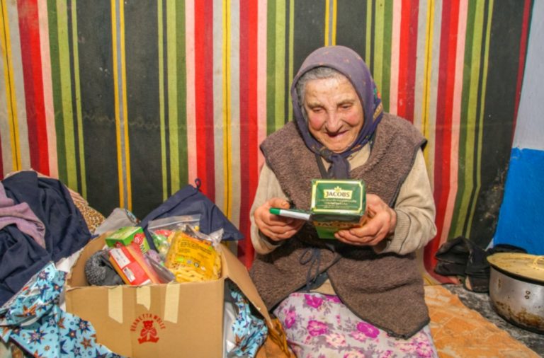 Une grand-mère découvre les objets d'un carton à côté d'elle. Elle est assise sur un lit, les cheveux couverts par un châle. Derrière, un papier peint aux rayures multicolores