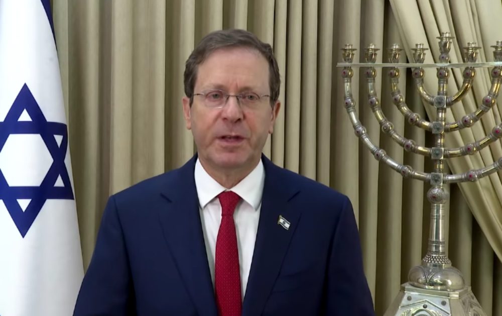 Isaac Herzog, 61 ans, parle vers l'objectif/ Il porte des lunettes et un costume bleu foncé. Derrière lui, le drapeau israélien et un candélabre à neuf branches.