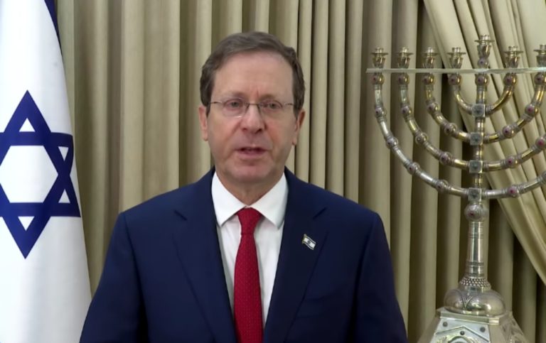 Isaac Herzog, 61 ans, parle vers l'objectif/ Il porte des lunettes et un costume bleu foncé. Derrière lui, le drapeau israélien et un candélabre à neuf branches.