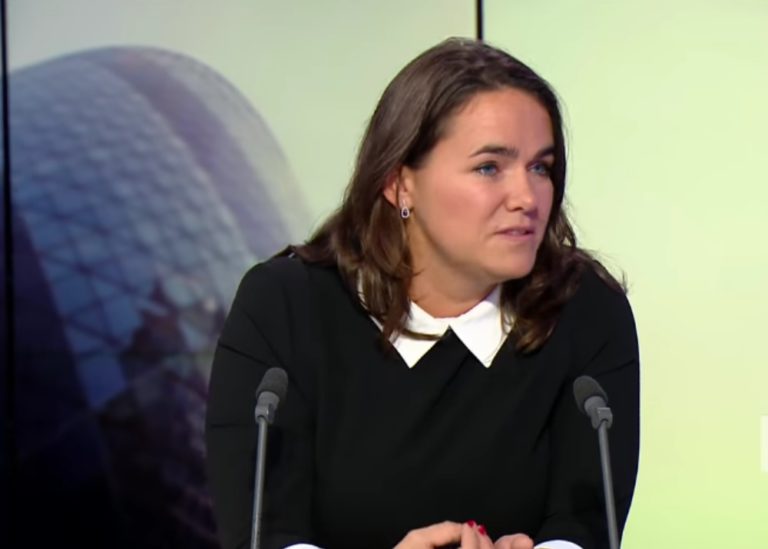 Katalin Novák, jeune femme brune aux yeux bleus, parle sur un plateau télé, le buste légèrement penché sur une table. Elle porte un pull over noir. Devant elle, deux micros fins.