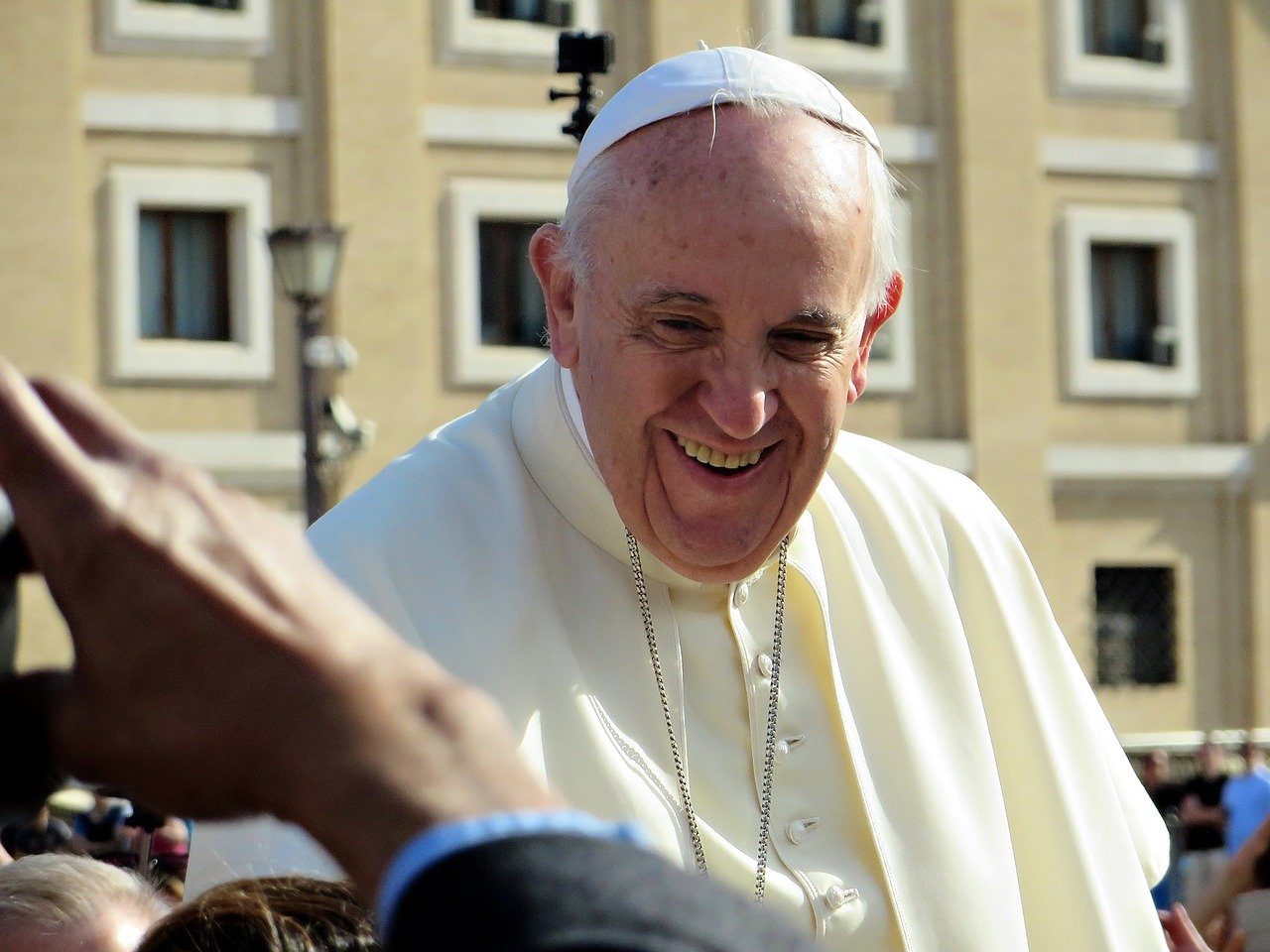 Le pape François sourit devant l'objectif d'un journaliste. il est vêtu de blanc. On voit son buste, au-dessus de la main du photographe qui est hors champs.