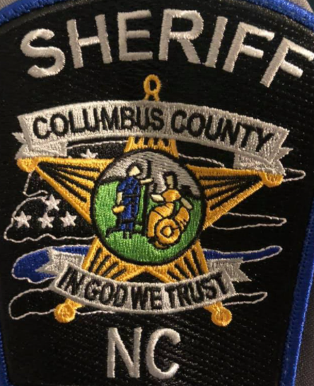 Gros plan sur un insigne de police; On peut y lire: "Sheriff - Columbus county - In God we trust. Les mots sont brodés autour autour d'une étoile jaune sur fond noir. Au centre, une scène champêtre avec deux petits personnages.
