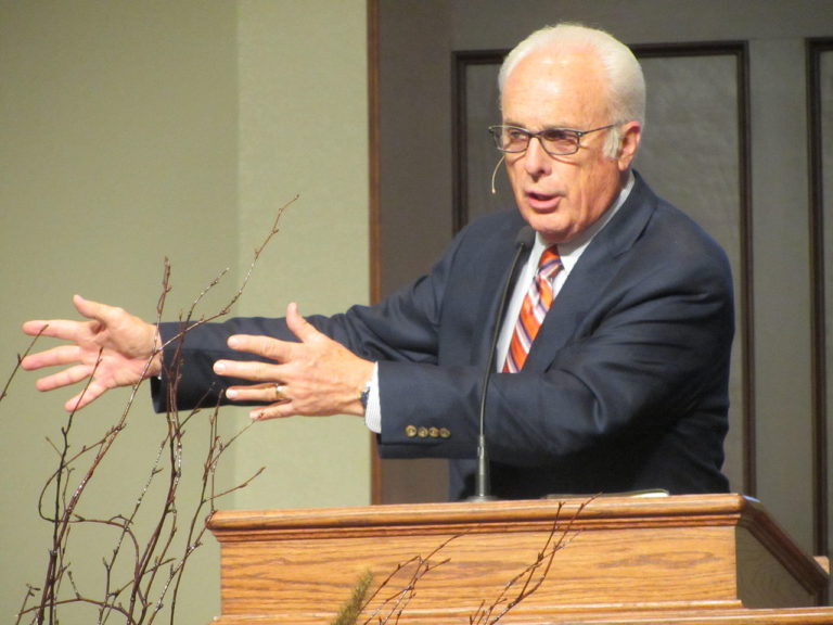 John MacArthur, environ 70 ans, prêche derrière un pupitre. Concentré, il explique quelque chose, les bras étendus vers sa droite, mains ouvertes.