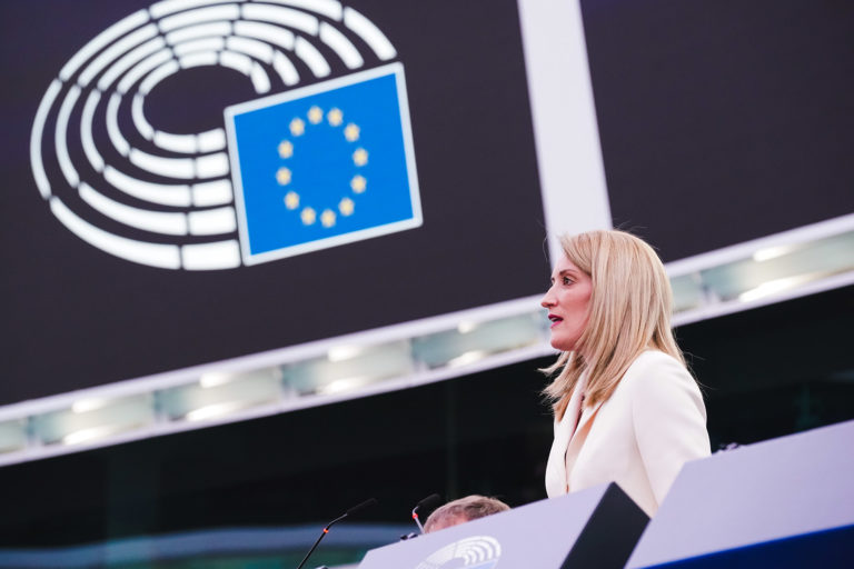 Roberta Metsola durant une conférence de presse. Elle est de profil et derrière elle, on voit le symbole étoilé de l'Union européenne. Elle est blonde et porte une veste blanche.