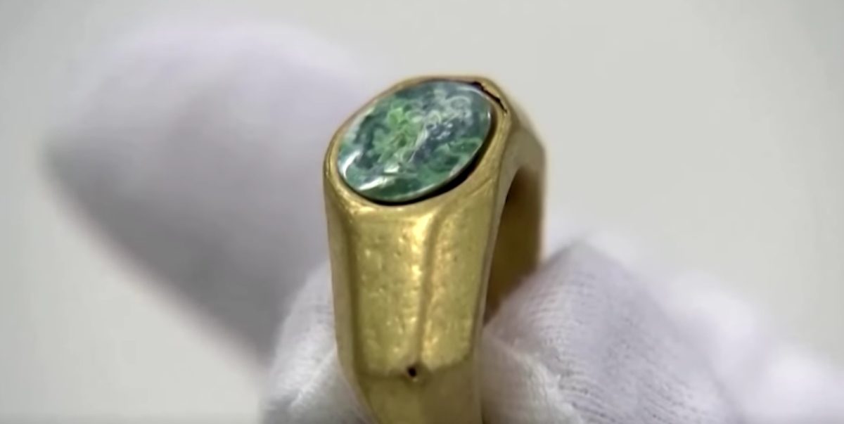 De près, une bague dorée sertie d'une pierre ronde plate aux couleurs vertes. Des doigts gantés de blanc la tiennent.