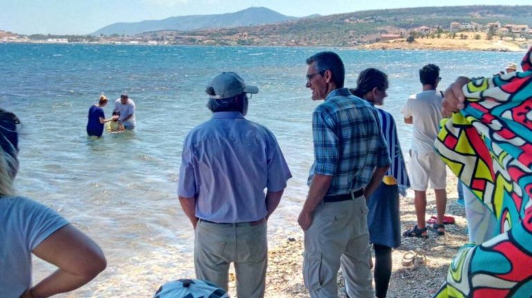 Deux personnes en baptisent une autre dans un lac. Au premier plan sur la rive, des hommes, les mains dans les poches, discutent entre eux.