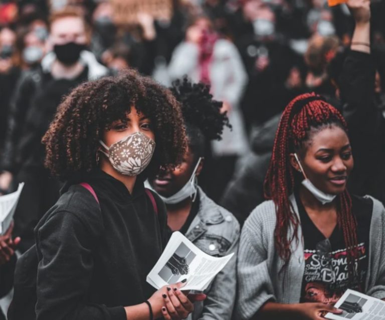 Une foule de gens portant des masques anti-covid. Plusieurs tiennent un imprimé dans leurs mains. Au premier plan, une jeune fille d'origine africaine regarde vers l'objectif.