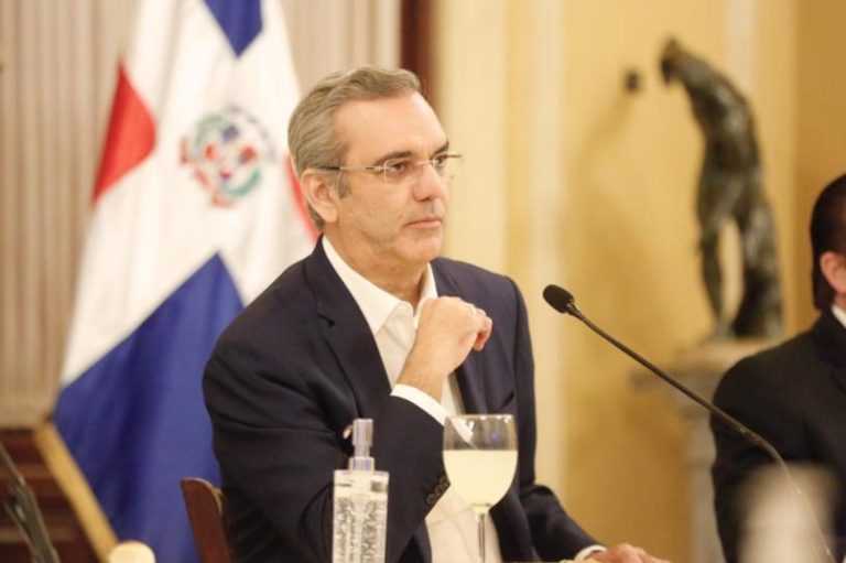 Luis Abinader Corona, la cinquantaine, a le coude posé sur une table, sur laquelle on voit un verre à pied. La main pliée, il semble réfléchir aux propos d'une personne hors-champ. Il porte des lunette et un costume sombre. Derrière lui se trouve un drapeau de la République dominicaine.