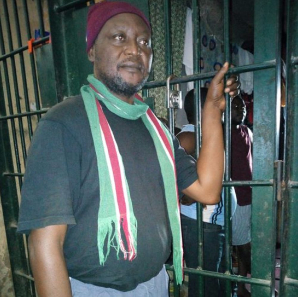 Luka Binniyat, nigérian, tient de sa main gauche une grosse grille, derrière laquelle on aperçoit plusieurs personnes. Il regarde vers la droite. Il porte un tee-shirt noir et une écharpe fine.