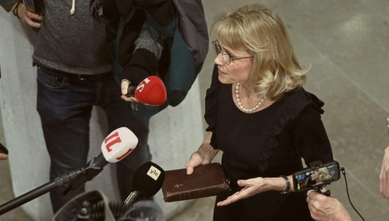 Païvi Räsänen, femme blonde habillée en noir, parle devant plusieurs micro tendus devant elle. Elle tient une Bible entre ses mains.