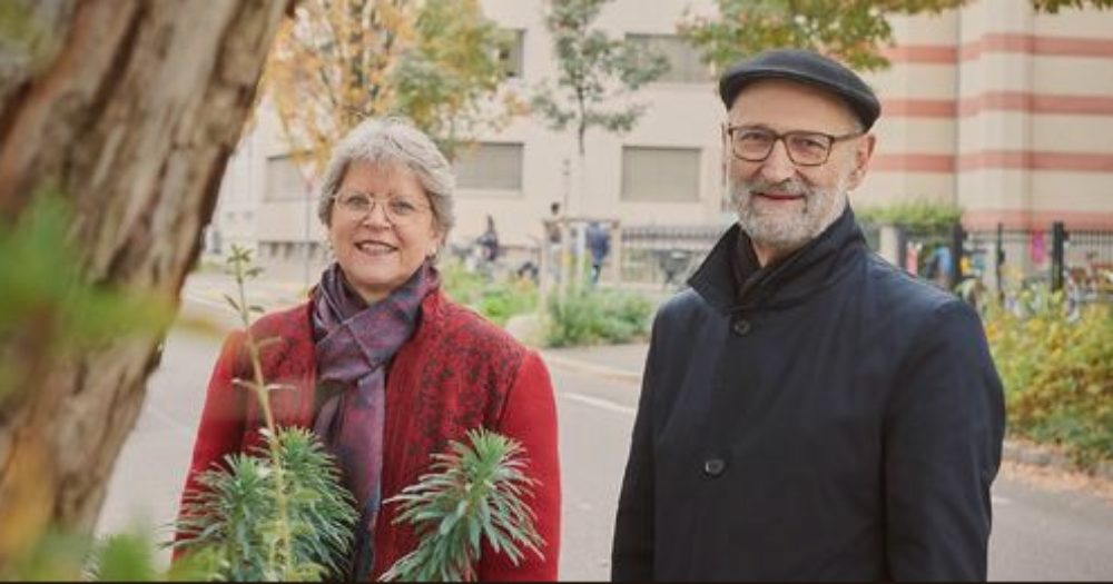Rita Famos, en manteau rouge, et Ralph Lewin, en noir, sont dans un parc et regardent vers l'objectif en souriant. Ils portent tous deux des lunettes et semblent heureux.