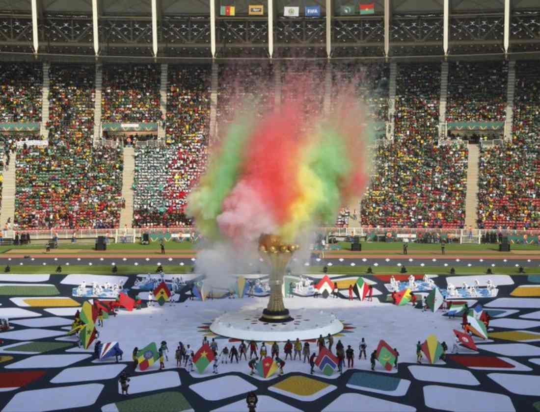 Cérémonie d'ouverture de la Coupe d'Afrique des Nations. Une grosse gerbe de poudres multicolores s'élève au-dessus d'une immense colonne évasée représentant une coupe d'or. Tout autour, des drapeaux et des gens regardent. Ils sont minuscules à côté de cette construction.