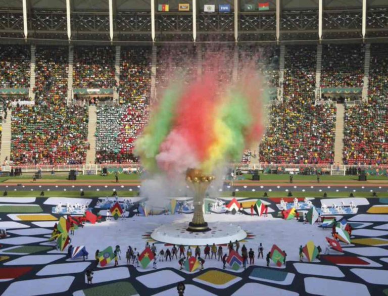 Cérémonie d'ouverture de la Coupe d'Afrique des Nations. Une grosse gerbe de poudres multicolores s'élève au-dessus d'une immense colonne évasée représentant une coupe d'or. Tout autour, des drapeaux et des gens regardent. Ils sont minuscules à côté de cette construction.