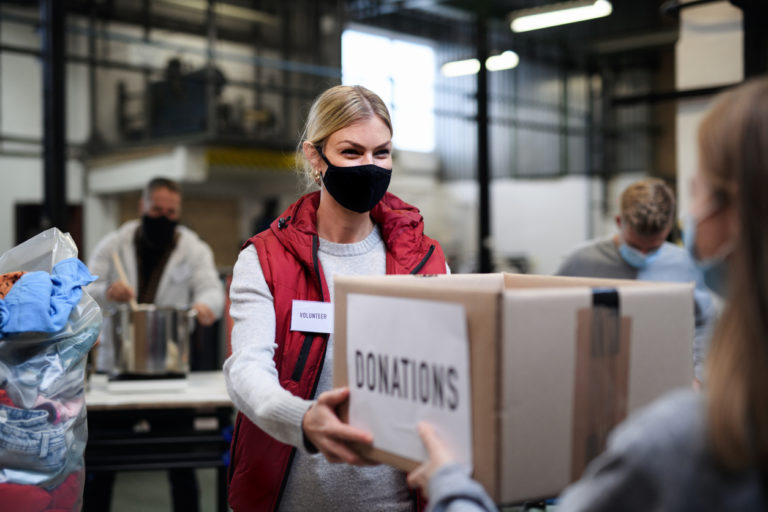 Dans un entrepôt animé, une jeune femme blonde tend un gros colis marque "donation" à une personne en face d'elle. Elle porte un masque anti-covid.