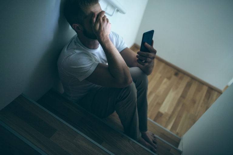 Un homme jeune est assis au pied d'un escalier en bois en intérieur. Il couvre ses yeux avec sa main crispée. Il tient face à lui un smartphone et porte a priori un pyjama. il est dans la pénombre.
