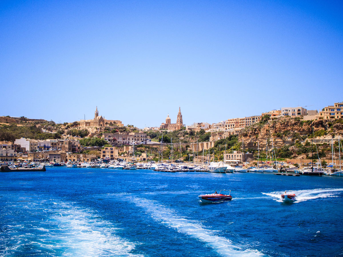 Vue d'une côte de l'île de Malte depuis la mer. Il y a un grand ciel bleu. Sur la côte, une multitude de petits bâtiments et de maisons.
