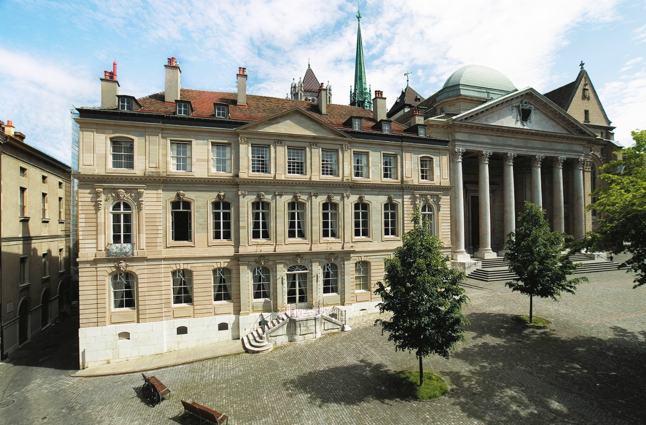 Photo du musée de la Réforme à Genève. C'est un gros bâtiment rectangle à deux étages en pierre ocre, avec de nombreuses moulures géométriques. Certaine représentent aussi des visage. La photo est prise depuis un point haut placé. On voit une cour avec un petit arbre devant.