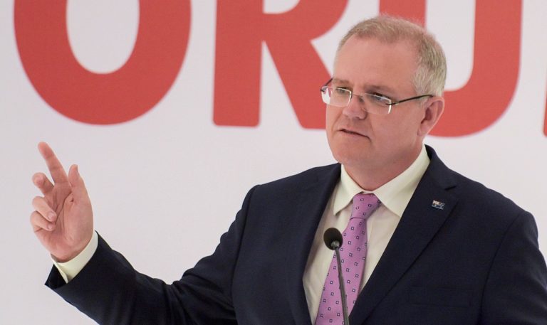 Photo de buste de Scott Morrison, la cinquantaine grisonnante. Il fait un discours, la main droite levée. Il porte un costume sombre et des lunettes. Il a l'air préoccupé.