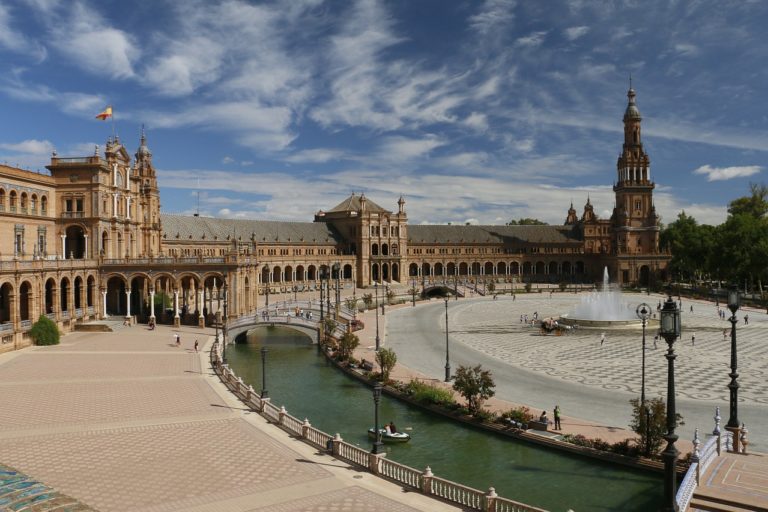 Vue d'une immense place en forme de cercle à Séville, avec une fontaine au milieu. Des bâtiments historiques longent la place.